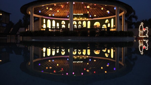 A circular building with a thatched roof, illuminated by colorful lights, reflecting in a pool. - PRAKARAM RESORT AND CAFE (Ramnagar)