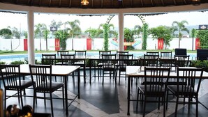A covered outdoor dining area with tables and chairs, a pool in the background, and a view of greenery and a building with a sign. - PRAKARAM RESORT AND CAFE (Ramnagar)