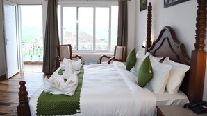 A hotel room with a large bed, two chairs, a view of the outdoors, and a painting on the wall. - PRAKARAM RESORT AND CAFE (Ramnagar)