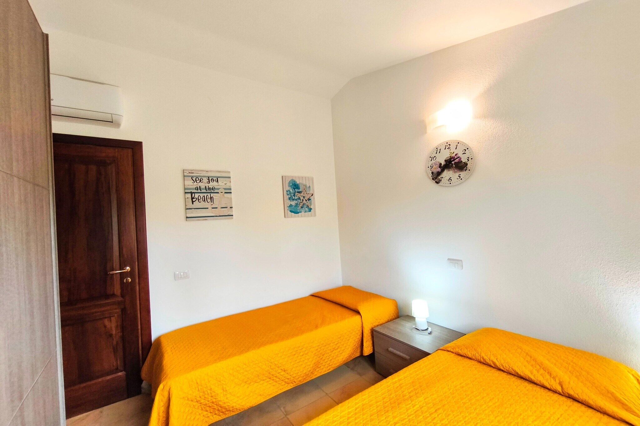Une chambre avec deux lits, une porte en bois, une horloge murale et un luminaire fixé au mur.