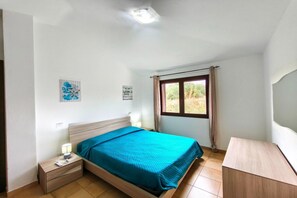 Quarto com cama, mesa de cabeceira, abajur e janela com cortinas.