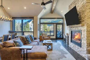 A spacious living room with a fireplace, large windows, and a flat-screen TV mounted on the wall. - Fireplace, Game Room & Mountain Views | Near Golf & Dining | Keystone River Run (Dillon)