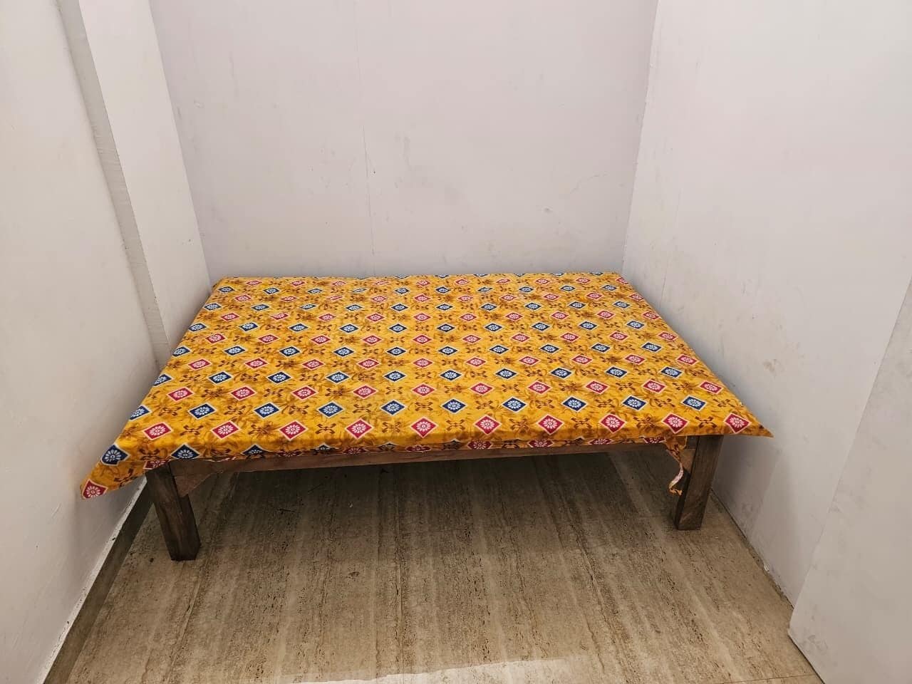 A small wooden table with a yellow patterned tablecloth in a corner of a room with plain white walls and a wooden floor.