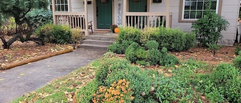 Una casa con un porche acogedor, una puerta verde y un jardín bien cuidado con diversas plantas y un camino que lleva a la entrada.