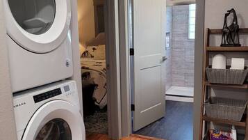 A laundry room with two stacked washing machines, a wooden floor, a rug, and a door leading to a bedroom.