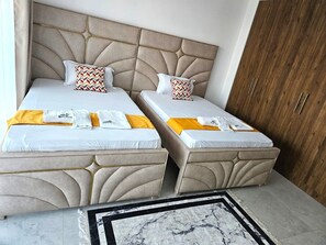 A hotel room with two beds, each with a beige headboard, white linens, and patterned pillows. There are towels and a small rug on the floor. - Modern 2BR with Pool Near Beach & City Mall, Nyali (Mombasa)