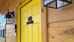Une porte jaune ornée d’une enseigne d’accueil en forme d’ours, une lanterne noire et un mur en bois.