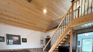 Un vaste espace de vie doté d’un plafond en bois, d’un canapé en cuir marron, d’une table ronde en bois avec des tabourets et d’un escalier menant à un niveau supérieur.