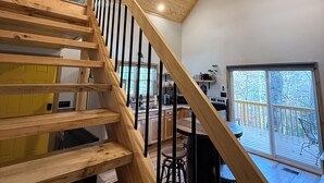 Un escalier en bois, surmonté d’un ventilateur de plafond, mène à une cuisine dotée d’une grande fenêtre et donnant sur une terrasse.