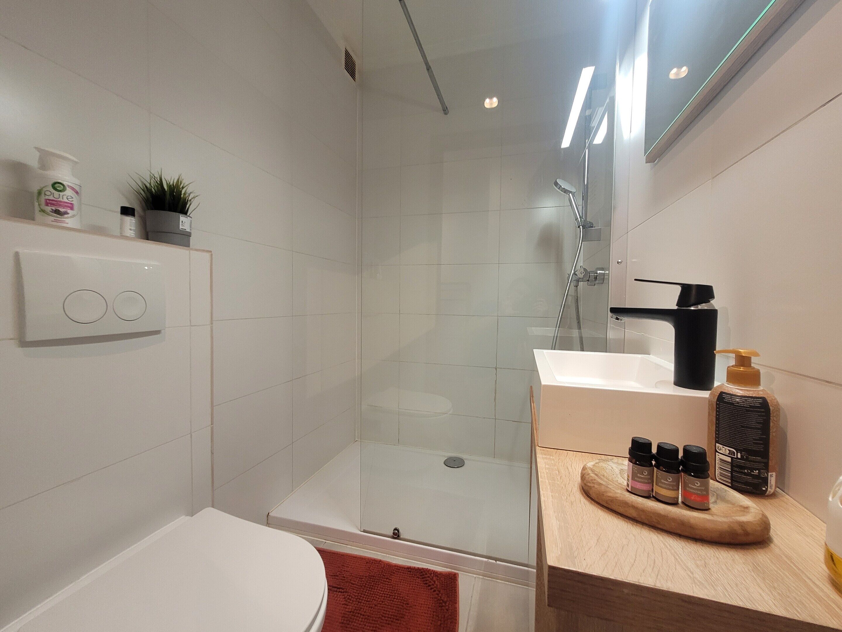 Une salle de bain moderne avec une douche à parois de verre, des toilettes blanches et un meuble-lavabo en bois où sont rangés les articles de toilette.
