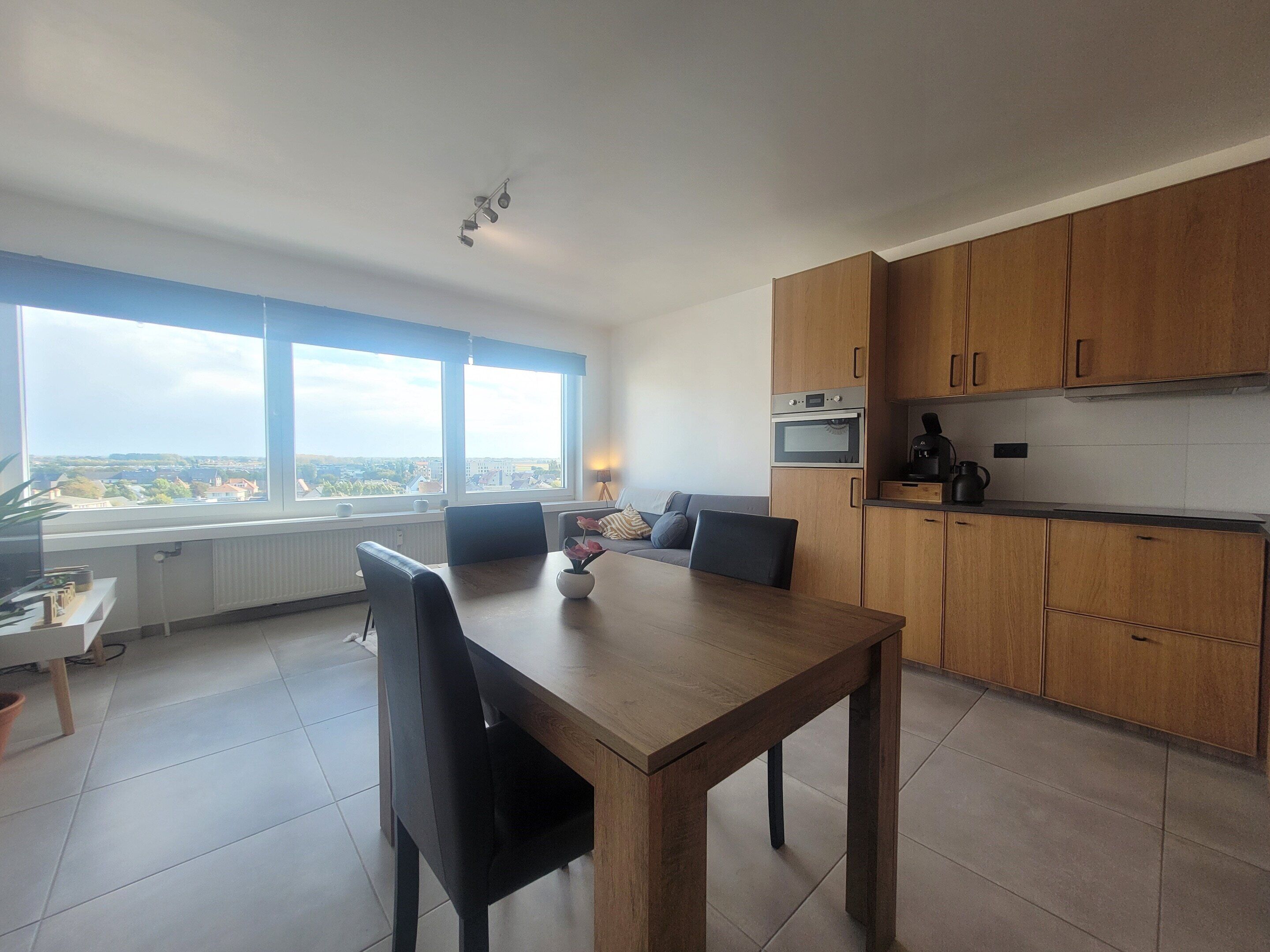 A modern kitchen with wooden cabinets, a dining table, and a view of the city through large windows.