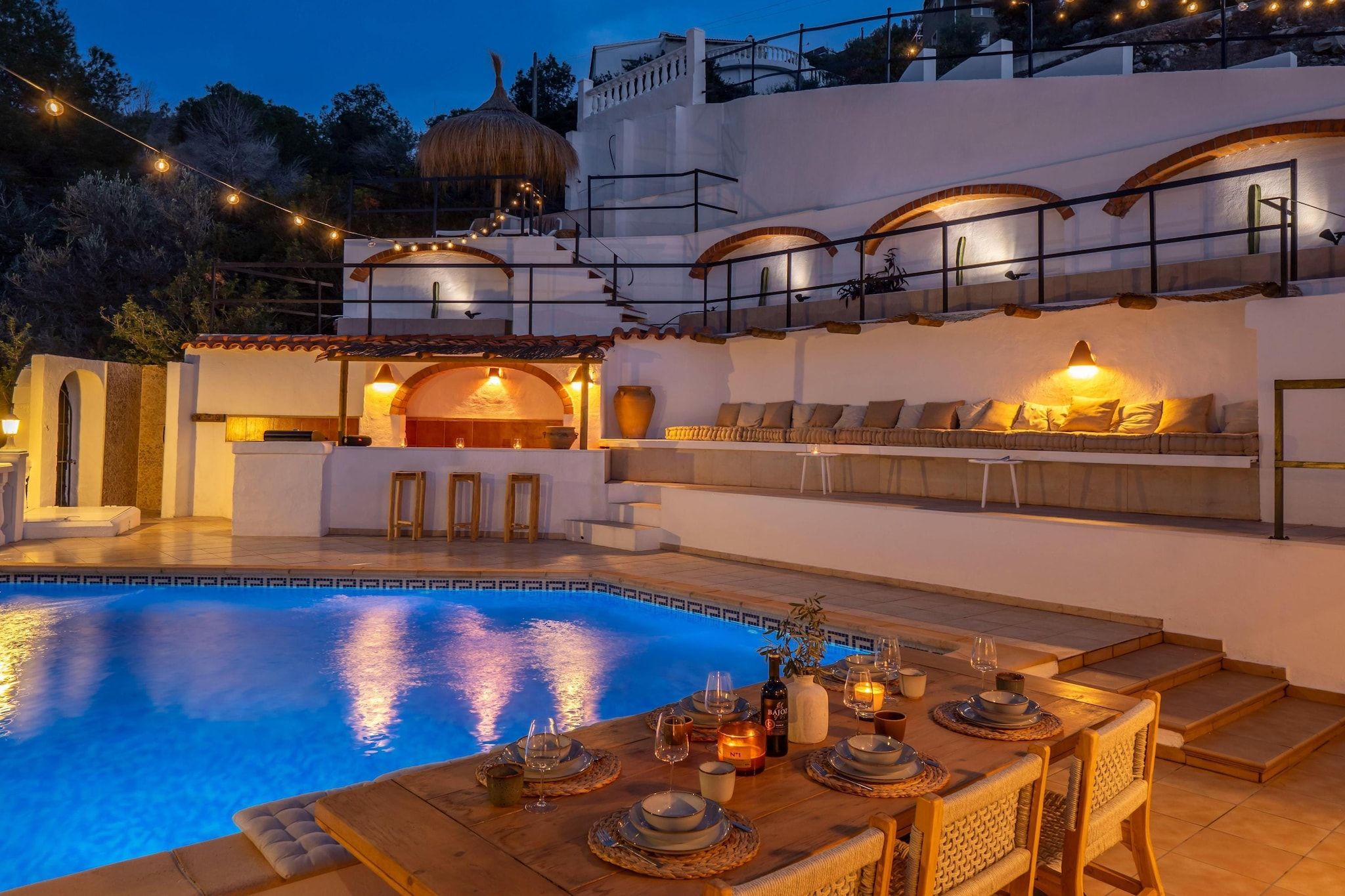 Un espace repas aménagé au bord de la piscine, avec une table dressée pour deux personnes, un coin salon avec des coussins et une vue sur la piscine.