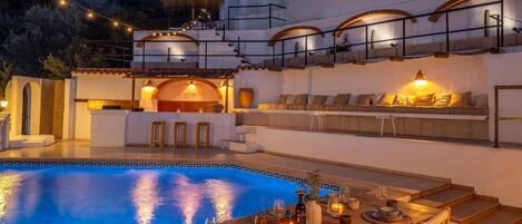 A poolside dining area with a table set for two, a seating area with cushions, and a view of the pool.
