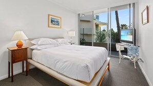 A hotel room with a bed, a TV, a desk with a chair, a window with a view of palm trees, and a painting on the wall. - Gorgeous 2-Bed Unit near Riverside Parks (Chiswick)