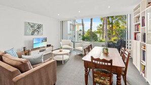 A modern living room with a large TV, comfortable seating, a dining area, and a bookshelf. - Gorgeous 2-Bed Unit near Riverside Parks (Chiswick)