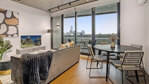 A modern living room with a large window, a sofa, a dining table, and a TV. - Modern, Executive 1 Bedroom Apartment With Balcony (Cremorne)