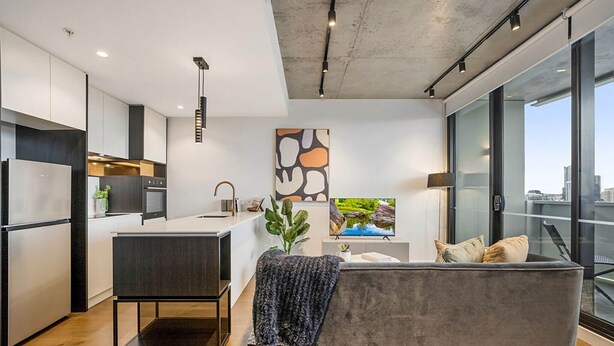A modern kitchen with a central island, a dining area with a grey sofa, and a large window offering a city view. - Modern, Executive 1 Bedroom Apartment With Balcony (Cremorne)