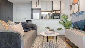 A modern living room with a sofa, a coffee table, and a TV. There is a kitchen area in the background with cabinets and appliances. - Modern, Executive 1 Bedroom Apartment With Balcony (Cremorne)