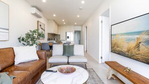 Una sala moderna con un sofá de cuero marrón, un sofá blanco y una mesa de centro de mármol. En el fondo, hay un comedor con una mesa y sillas.