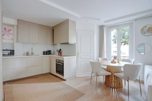 Eine moderne Küche mit weißen Schränken, ein Essbereich mit einem runden Holztisch und weißen Stühlen sowie ein großes Fenster mit Vorhängen.