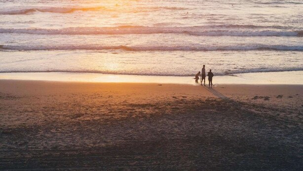 Une scène de plage avec un coucher de soleil, des vagues et trois personnes qui se promènent le long du rivage.