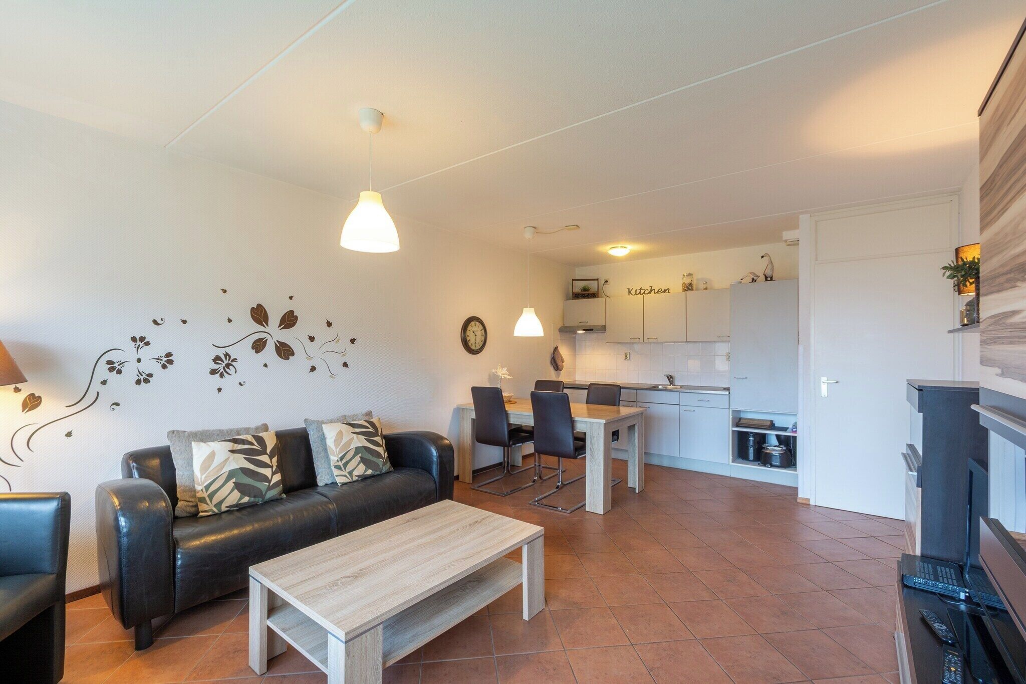 Un salon moderne avec un canapé en cuir noir, une table basse en bois et un coin repas avec une table et des chaises blanches. On aperçoit une cuisine à l’arrière-plan, avec des meubles blancs et une enseigne indiquant « Cuisine ».
