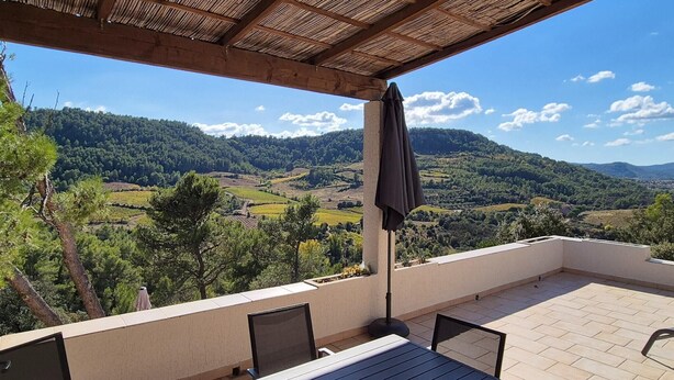 Een terras met uitzicht op een heuvelachtig landschap, een tafel, stoelen en een pergola met een parasol.
