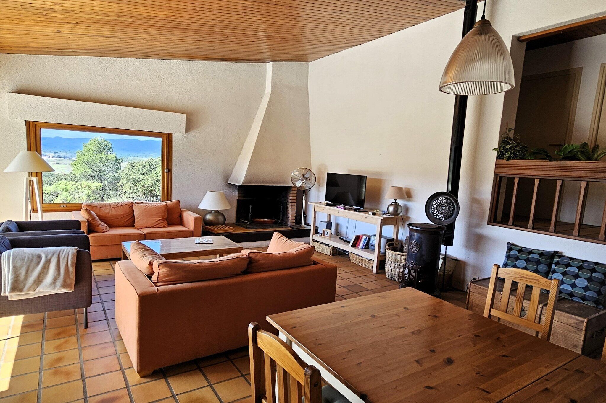 A cozy living room with a wooden ceiling, a large window with a scenic view, a fireplace, a TV, and a dining area with a wooden table and chairs.