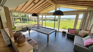 Une pièce spacieuse équipée d’une table de billard, d’un canapé et de grandes fenêtres donnant sur un paysage verdoyant.