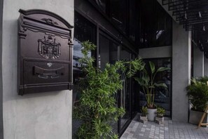 Una entrada de edificio con un buzón metálico, plantas en macetas y pavimento de baldosas.