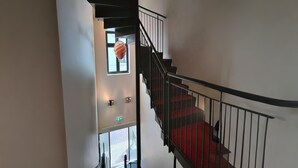 Un escalier avec des rampes noires et des marches recouvertes d’une moquette rouge, menant à un palier où est suspendu un luminaire.