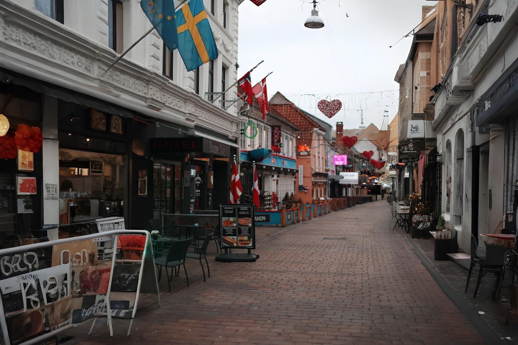 En brostensbelagt gade med butikker og restauranter, udsmykket med flag og hjerteformede dekorationer.
