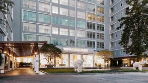 A modern hotel with a covered entrance, named "The Platinum Suite," featuring a large glass facade and a central courtyard with a statue. - Platinum Luxury Suite | Modern City Getaway (Bangkok)