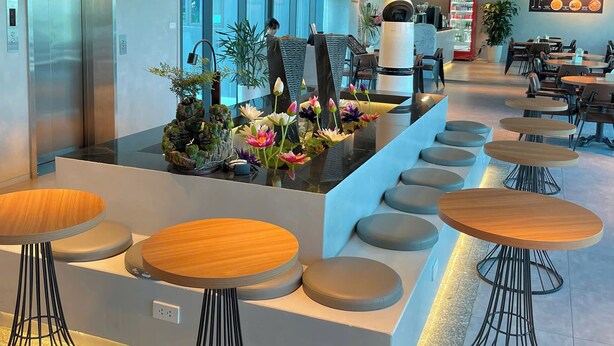 Un espace intérieur moderne doté d’un coin salon central avec des tables rondes et des tabourets, entouré de verdure et agrémenté d’un menu fixé au mur.