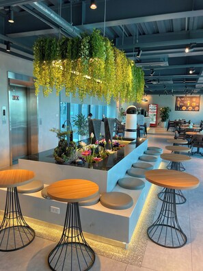 A modern indoor space with a central seating area featuring round tables and stools, surrounded by greenery and a wall-mounted menu. - Uberzen Residence Hotel (Hai Phong)