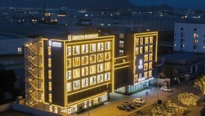 Un hôtel moderne, reconnaissable à son grand panneau « UBERZEN RESIDENCE », doté d’une cour intérieure avec un espace de stationnement et offrant une vue sur le paysage urbain environnant.