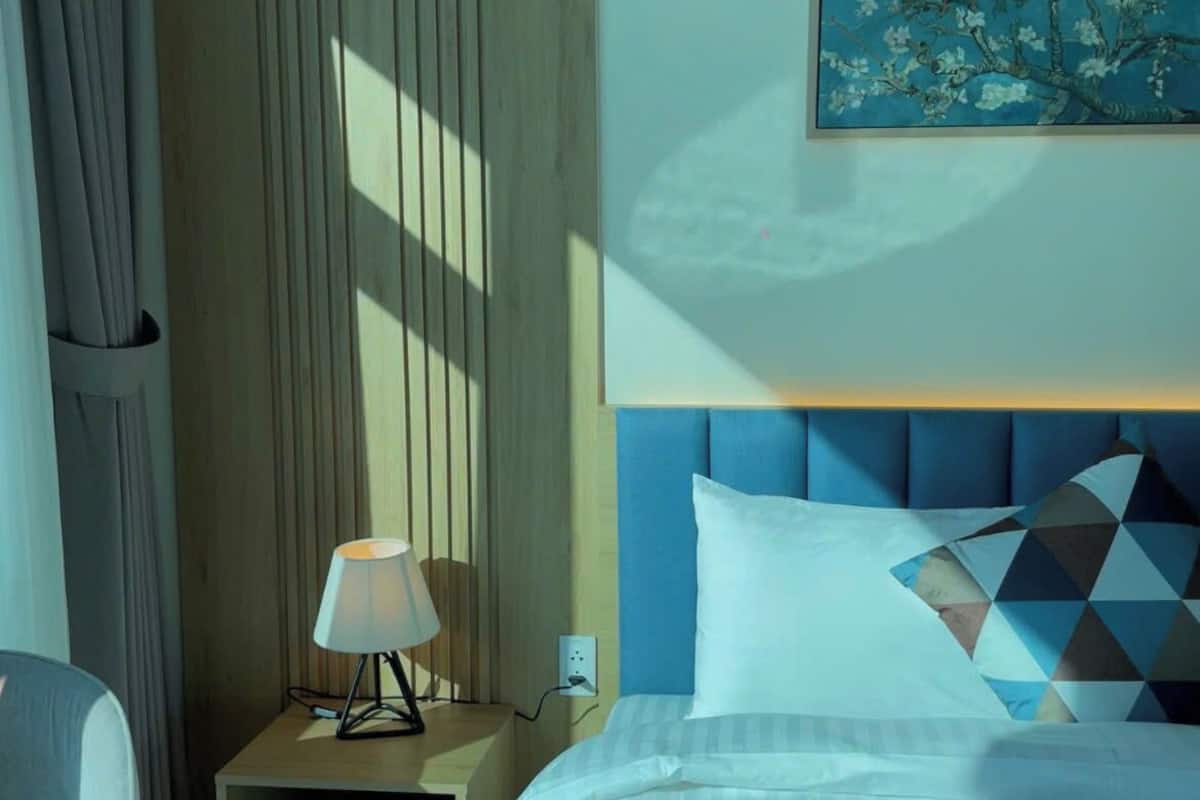 Habitación de hotel con una cama, una mesita de noche con lámpara, una silla y un cuadro en la pared.