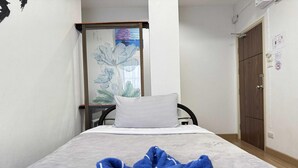 Een kleine hotelkamer met een bed, een wit kussen en een blauwe handdoek op het bed. Er is een houten deur en een witte muur met een raam.