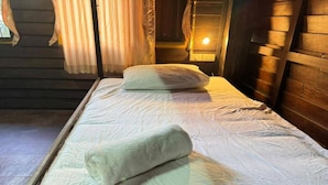 Ein Einzelbett mit weißer Matratze und Kopfkissen, ein aufgerolltes weißes Handtuch, Holzwände und ein Fenster mit Vorhängen.