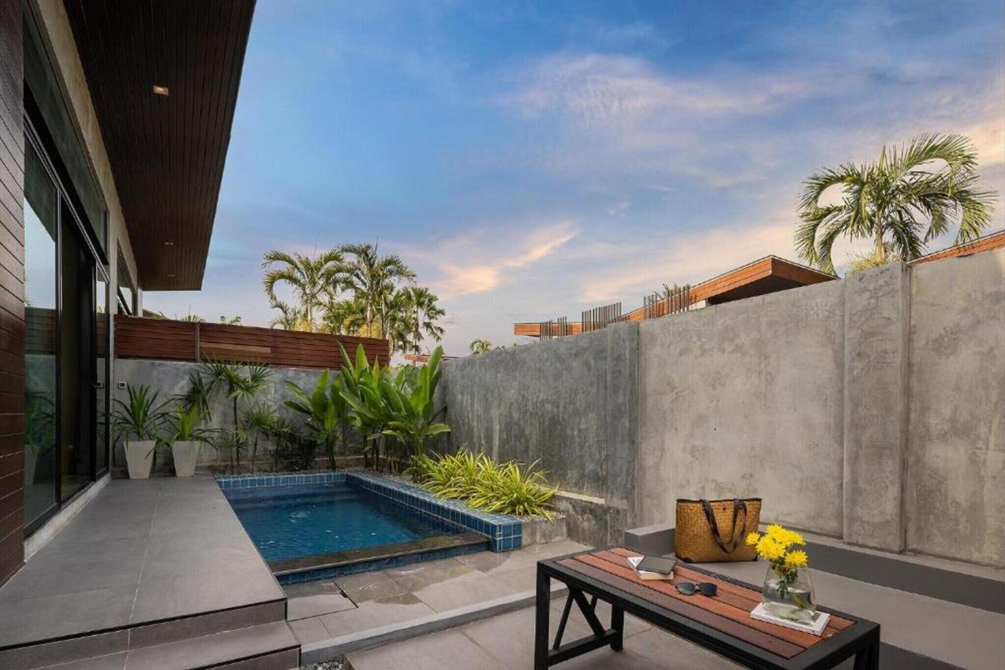 Un área de piscina moderna al aire libre con cubierta de madera, pared de concreto y plantas tropicales.