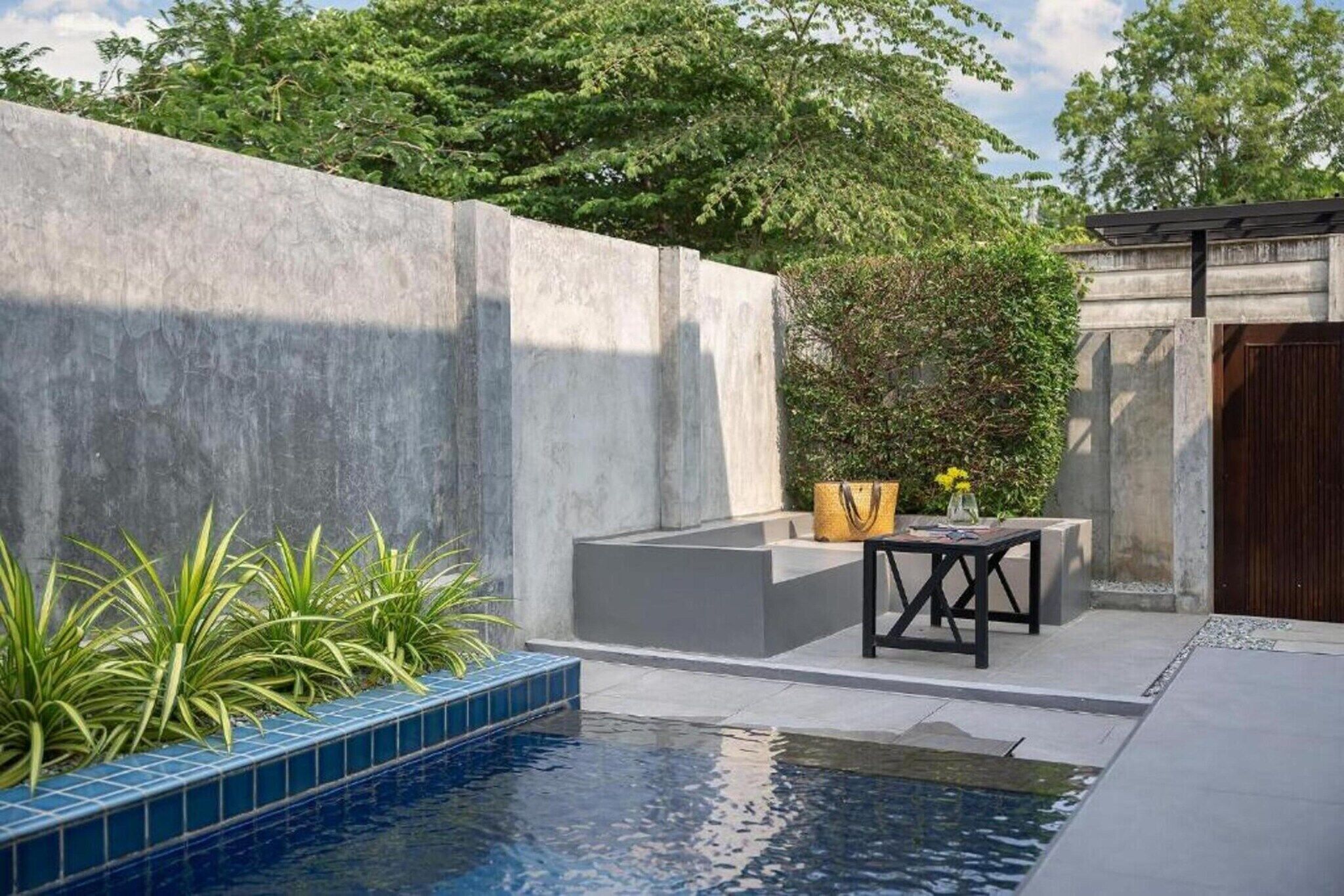 A modern outdoor pool area with a concrete wall, a small seating area with a table and chairs, and a potted plant.