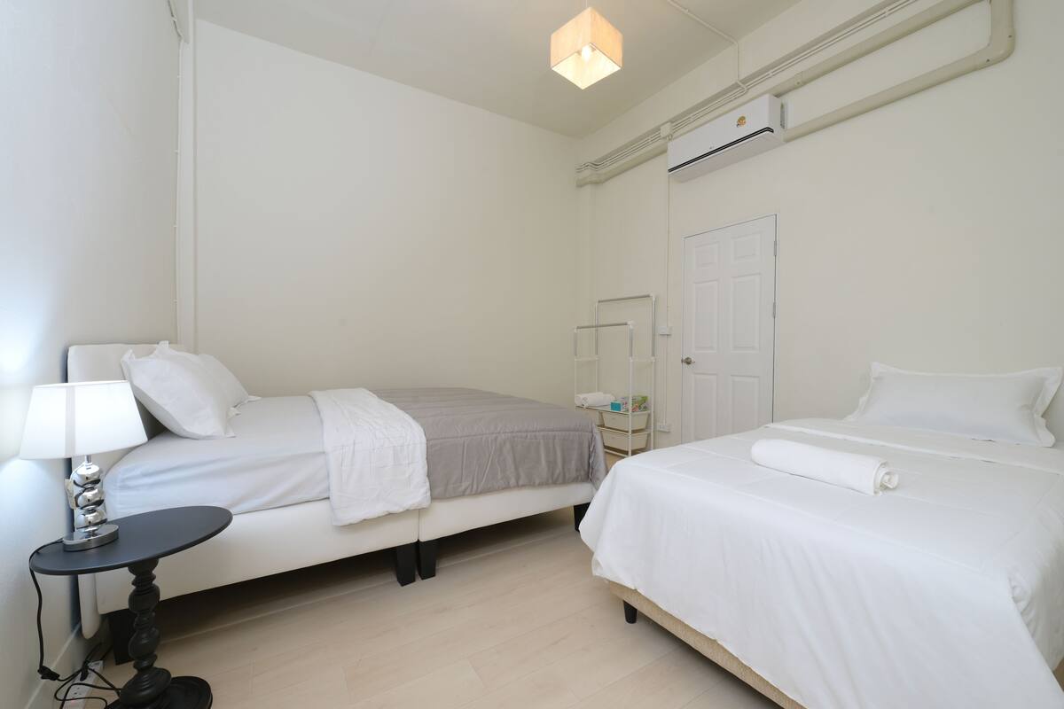 Ein Hotelzimmer mit zwei Betten, einem kleinen Tisch mit Lampe, einem Nachttisch, einem Kleiderschrank und einer Klimaanlage.