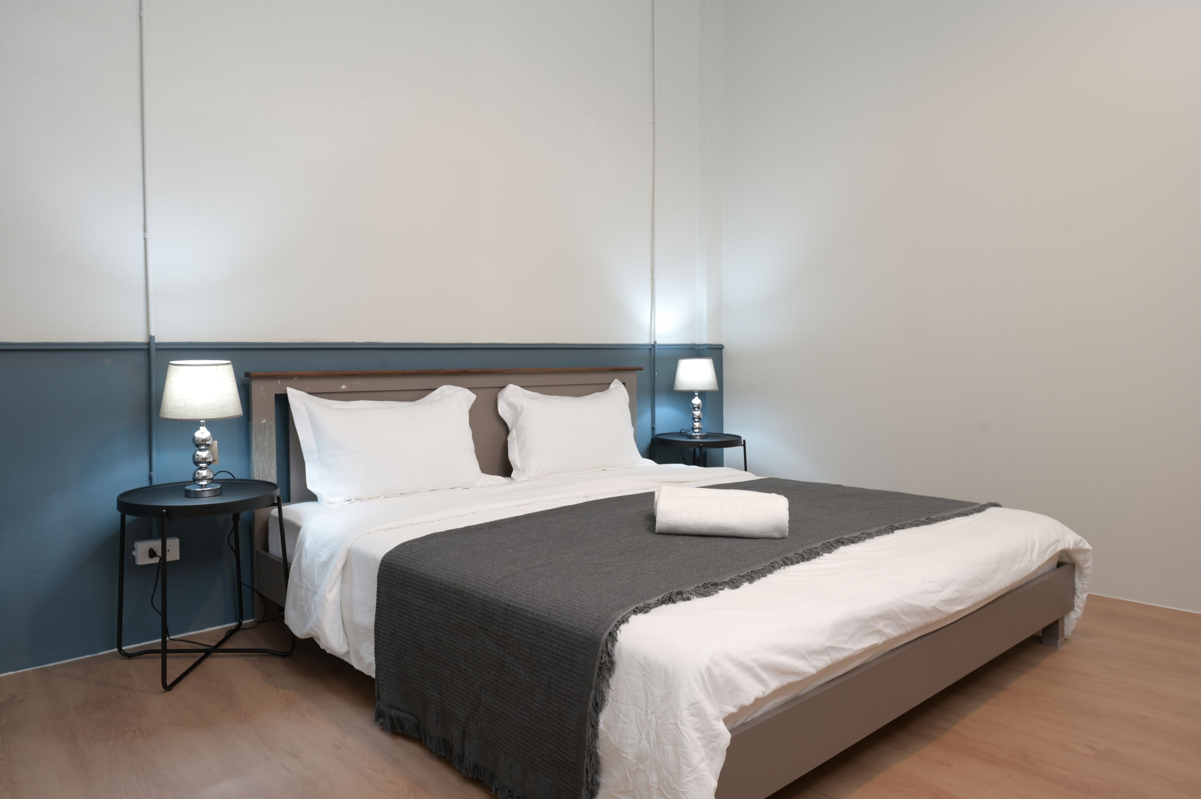 Cama bem arrumada com manta cinza, duas luminárias laterais e travesseiro branco sobre piso de madeira.