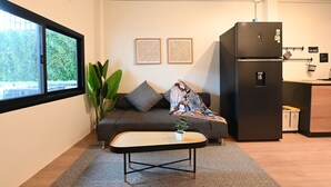 A compact living space with a sofa, a small table, a refrigerator, and a window with a view of greenery. - La Or Heritage Suites MRT Hua Lamphong (Bangkok)