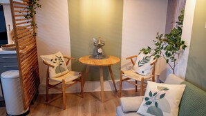 Una habitación acogedora con dos sillas de madera, una mesita redonda con un adorno, un sofá blanco con un cojín verde y una planta.
