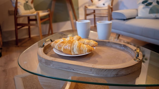 Una mesa con cubierta de vidrio y una bandeja de madera con croissants y dos tazas blancas.