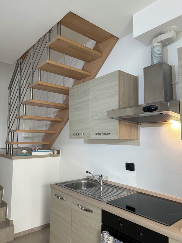 A modern kitchen with a wooden staircase, built-in cabinets, a stainless steel sink, and a black induction cooktop. - New standalone apartment 20 meters from the ocean (Santo Stefano al Mare)
