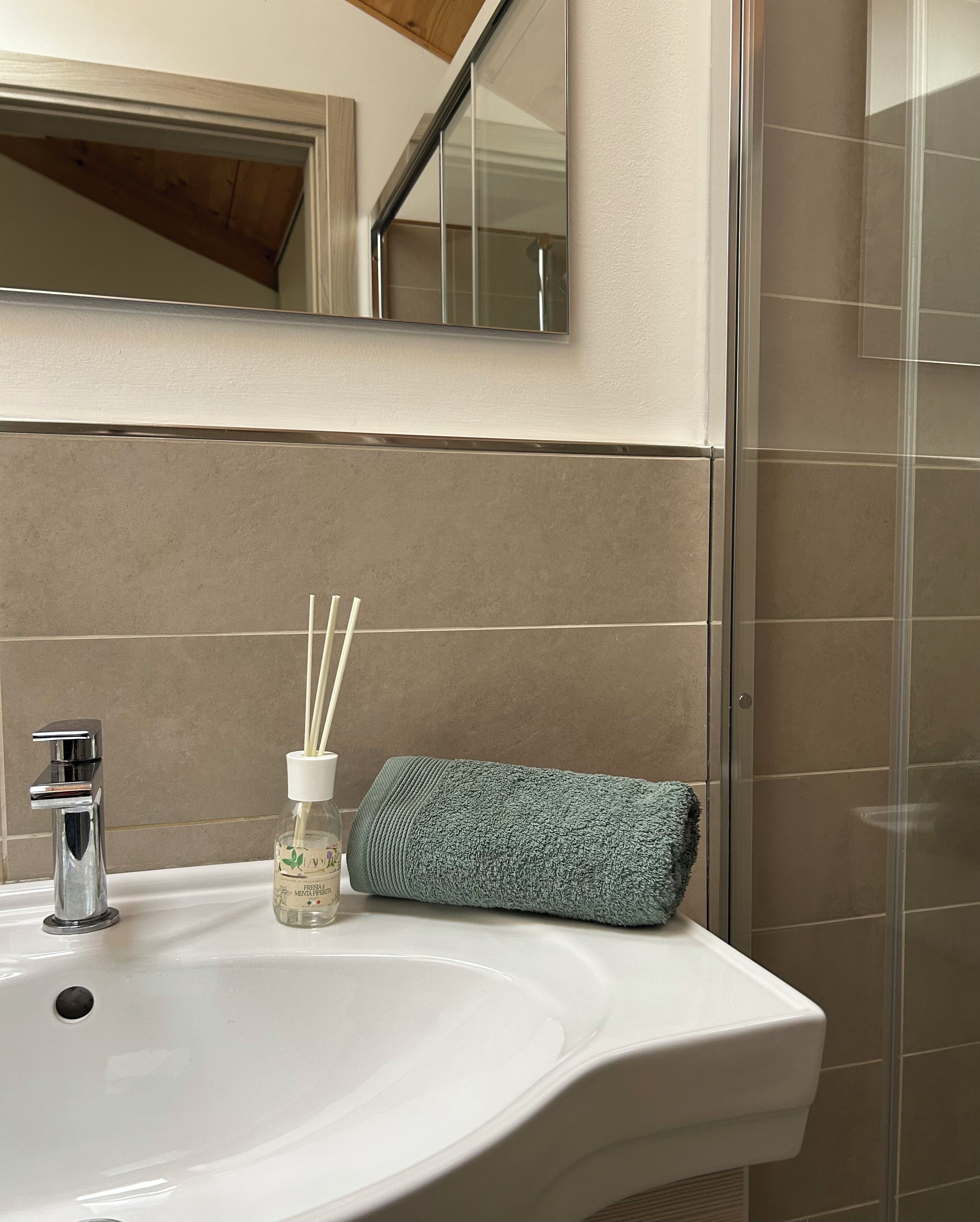A bathroom with a white sink, a mirror, a bottle of reed diffuser, and a rolled-up towel.