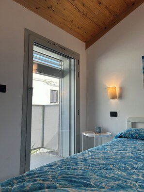 A bedroom with a bed, a small bedside table, a wall-mounted light, and a sliding glass door leading to an outdoor area. - New standalone apartment 20 meters from the ocean (Santo Stefano al Mare)