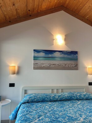 Um quarto com teto branco, vigas de madeira, cama com colcha azul estampada e uma pintura de uma paisagem praiana.